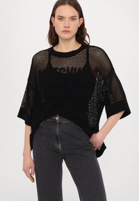 Melna oversized adīta džemperis ar sietiņa dizainu un īsām piedurknēm, ar apaļu apkakli un kontrastējošu tekstu, apvienots ar tumšām džinsām.