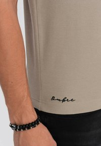 Béžová košile s krátkým rukávem z texturované látky, s černým vyšívaným logem blízko lemu. Černý korálkový náramek na zápěstí.