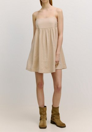 Kobieta ubrana w bezrękawnikową, beżową, luźną sukienkę mini, zestawioną z brązowymi zamszowymi kozakami do połowy łydki, na jednolitym tle.