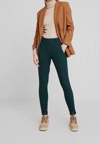Pantalons ajustés vert foncé avec une texture lisse, dotés d'une taille haute et de poches latérales, associés à un col roulé beige et un blazer marron.