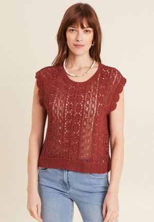 Haut en tricot couleur rouille avec des manches courtes à bords festonnés, caractérisé par une texture ajourée et un ourlet inférieur côtelé. Porté par-dessus un jean bleu.