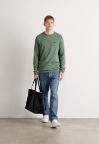 Green long-sleeve shirt, blue jeans, and white sneakers. Black tote bag with two handles is held by the wearer. Smooth texture, casual design.