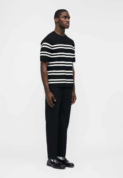 Άνδρας που ποζάρει φορώντας μαύρο κοντομάνικο πουκάμισο με λευκές οριζόντιες ρίγες, μαύρο παντελόνι, λευκές κάλτσες και μαύρα δερμάτινα παπούτσια μπροστά σε απλό φόντο.