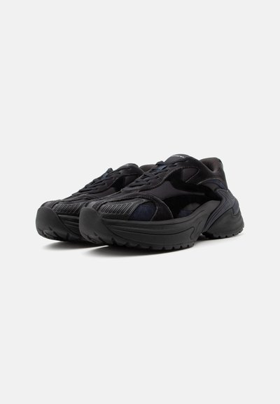 Paire de baskets de sport noires avec semelles épaisses, détails texturés et lacets, présentée en angle sur un fond clair uni.
