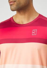 Kortermet treningsskjorte i røde og lyserosa striper, laget av pustende stoff med rund hals og en liten logo-detalj.