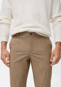 Beige trousers made of smooth fabric, featuring a flat front, belt loops, and a single button closure. Styled with a textured white sweater.