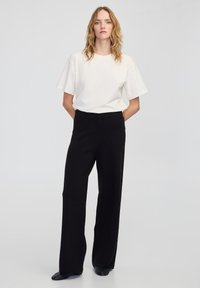 Weißes Kurzarm-T-Shirt in Kombination mit hochgeschnittenen, schwarzen Weitbeinhosen. Glattes Material, lockere Passform, minimalistisches Design.