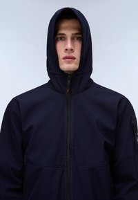 Młody mężczyzna w ciemnogranatowej kurtce z kapturem i zamkiem, zwrócony przodem na jednolitym jasnym tle.