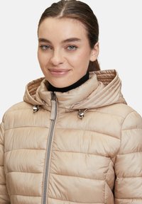 Beige Steppjacke mit strukturierter Oberfläche, Reißverschluss vorne und Kapuze. Merkmale sind silberne Hardware-Akzente am Kragen.