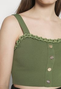 Top court côtelé vert avec larges bretelles, chaîne décorative le long de l'encolure et trois boutons dorés sur le devant.