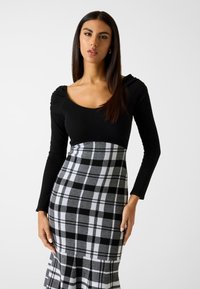 Schwarzes, geripptes Langarmoberteil mit runder Ausschnitt, kombiniert mit einem taillierten grauen Karorock mit ausgestelltem Saum und schwarzen Akzenten.