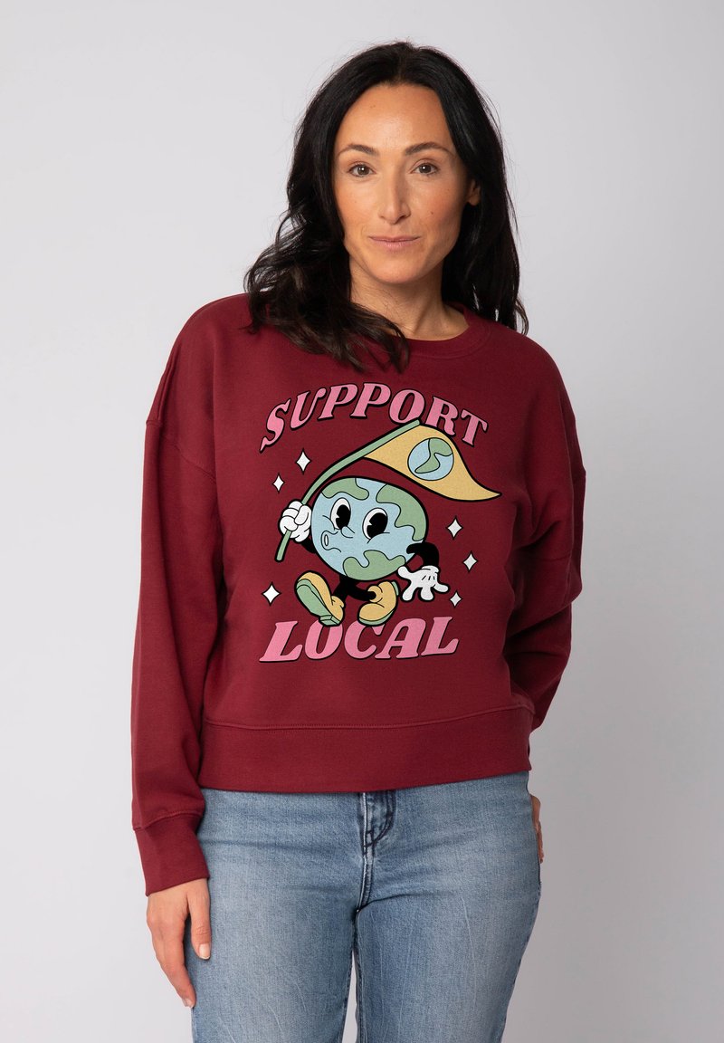 Bordeauxrode crewneck sweater met een grafisch ontwerp van een cartoonachtig Aarde-personage dat een vlag vasthoudt, met de tekst "SUPPORT LOCAL". Gemaakt van een katoenmix.