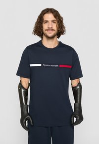 Dunkelblaues T-Shirt mit kurzen Ärmeln, weißen und roten horizontalen Streifen und dem zentrierten Logo "TOMMY HILFIGER". Schwarze Handschuhe und ein Prothesenarm sind sichtbar.