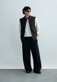 Zwarte mouwloze vest, witte sweatshirt en losse zwarte broek. Gestructureerde stof en een ontspannen pasvorm. Donkere zonnebril maakt de look compleet.