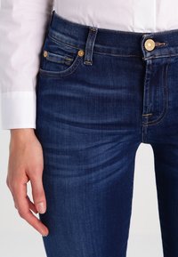 Dark blue denim jeans with a fitted design, featuring gold hardware accents, front pockets, and a smooth texture.
