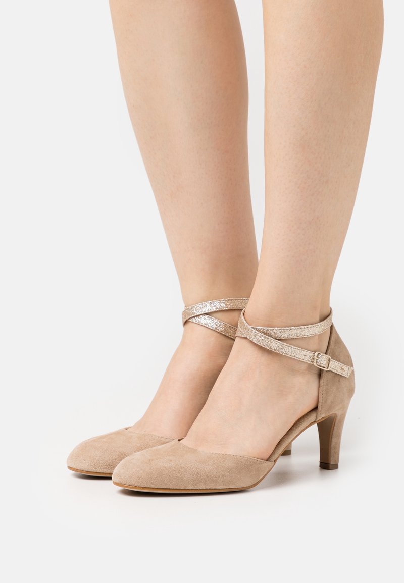 Chaussures en daim beige à talon moyen avec des brides croisées étincelantes autour de la cheville, attachées par de petites boucles sur pieds nus.