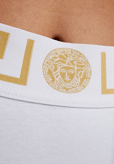 Vit elastisk midjeband med guldtexturerad Medusa-logotyp och mönstrad detaljer. Mjuk tyg med slät textur på kroppen.