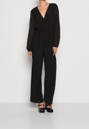 Kobieta ubrana w czarny kombinezon z długim rękawem, z przezroczystymi, pasiastymi rękawami i głębokim dekoltem w serek, w połączeniu z czarnymi lakierowanymi butami Mary Jane na obcasie.