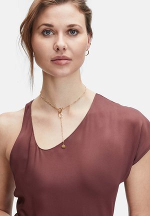 Goldkette mit rundem Anhänger, gestaffeltes Design. Das Model trägt ein burgunderfarbenes, leichtes Oberteil mit kurzen Ärmeln und rundem Ausschnitt.
