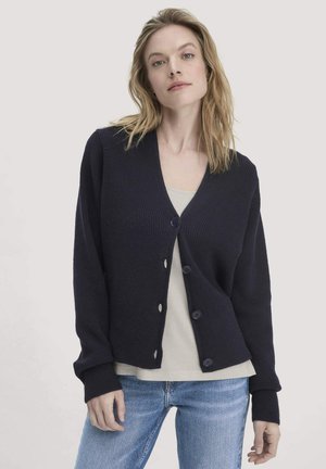 Cardigan a maglia blu navy con scollo a V, cinque bottoni e texture a coste. Maniche lunghe e vestibilità corta, abbinato a un top chiaro e jeans in denim.
