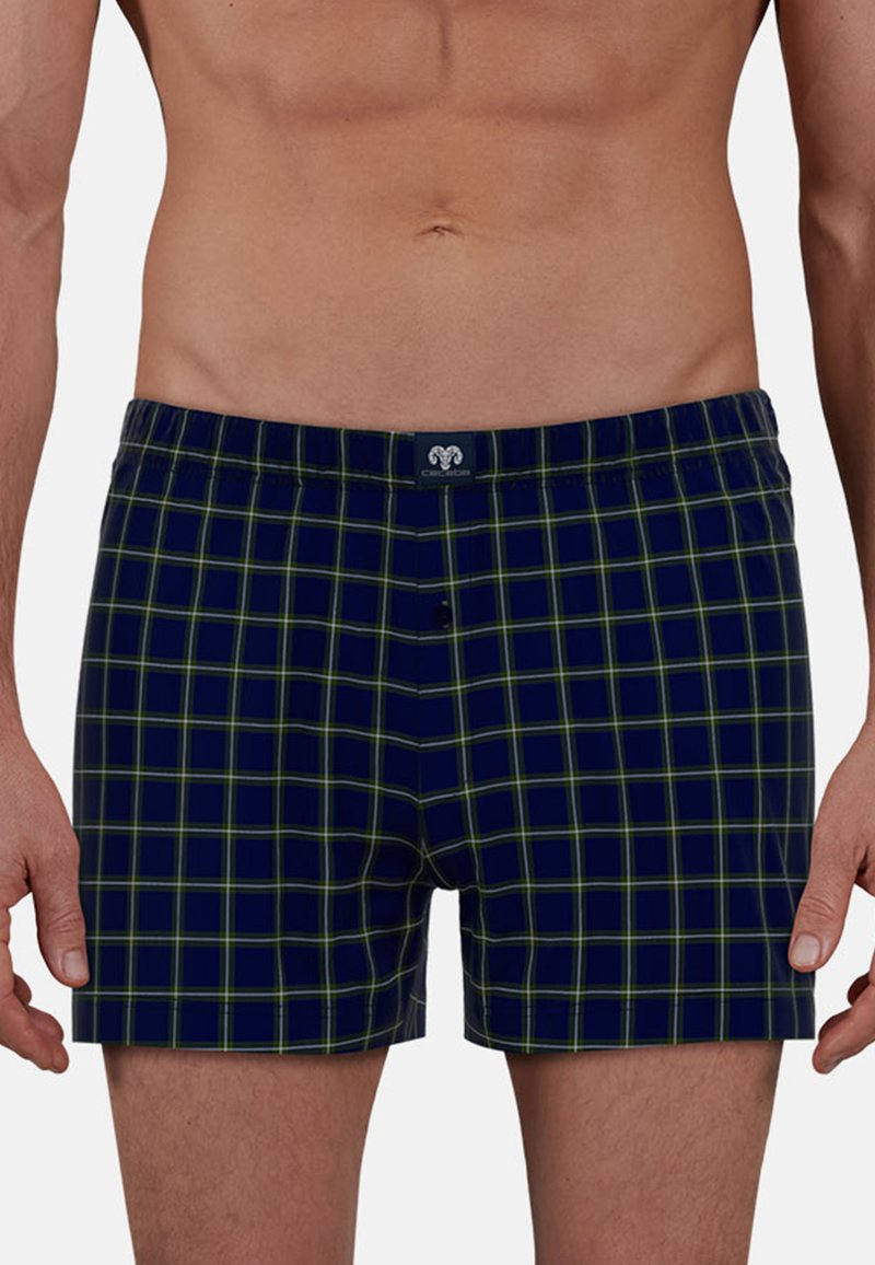 Marineblauwe boxershorts met een geel en groen geruit patroon, gedragen door een staande, naakte man tegen een effen achtergrond.