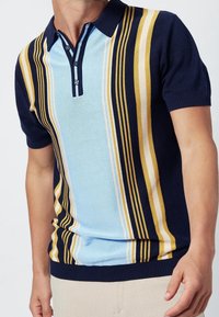 Man in een kortärmige polo met verticale strepen in marineblauw, lichtblauw, wit en mosterdgeel.