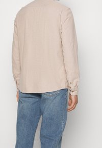 Beżowa koszula z długim rękawem i kołnierzykiem, widoczna faktura tkaniny oraz mankiety na guziki. W połączeniu z niebieskimi jeansami.