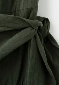 Material verde închis legat într-un nod formând o centură sau o eșarfă pe o haină, evidențiind textura și detaliile cusăturii.