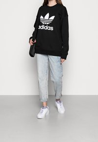 Sudadera negra con logo blanco de Adidas, jeans de denim azul claro y zapatillas blancas con acentos lavanda. Diseño sencillo y ajuste relajado.