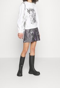 Vit sweatshirt med grafiskt tryck, metallisk silverkjol med lila blixtmönster, och svarta knähöga gummistövlar med texturerade sulor.