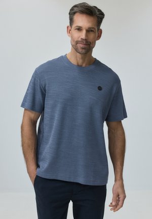 Mężczyzna o krótkich włosach i brodzie, ubrany w niebieską teksturowaną koszulę z krótkim rękawem i ciemne spodnie, stojący z jedną ręką w kieszeni.