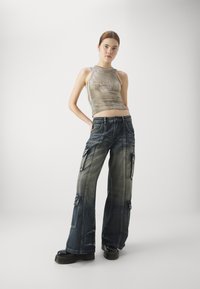 Cargo jeans v bledem modrem denimu s podloženimi žepi, v kombinaciji s svetlo bež kratkim topom. Manekenka pozira s rokami na bokih pred belo ozadje.