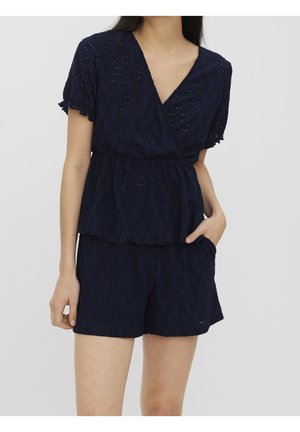 Kobieta ubrana w granatowy top z krótkim rękawem z ażurowej tkaniny z dekoltem w serek i pasujące szorty, z jedną ręką w kieszeni, na jednolitym tle.