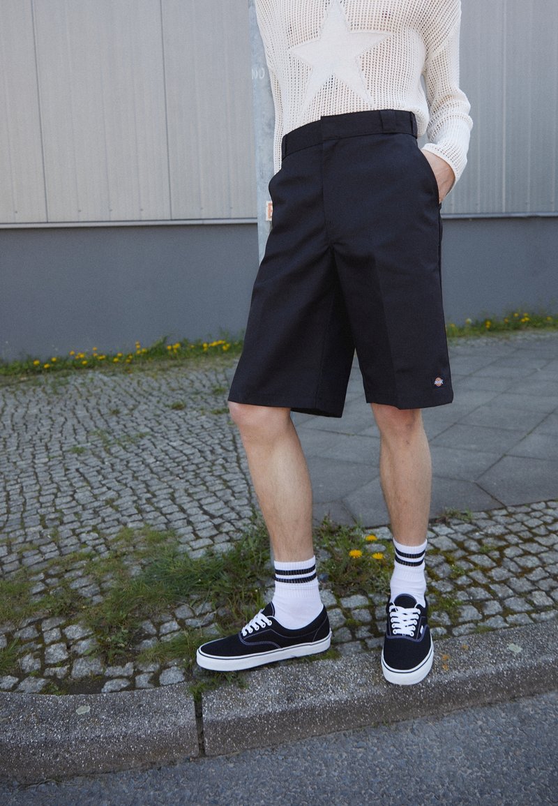 Personne portant un short noir aux genoux, des chaussettes blanches avec des rayures noires, des baskets noires et un pull blanc avec un motif étoile, debout sur le trottoir.