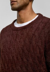 Garengebreide trui in donker bordeaux. Zacht, gestructureerd stof met een ronde halslijn, gedragen over een wit shirt.