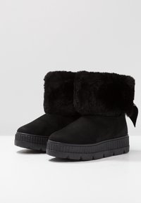 Svarta ankelstövlar med en fuskpälsfåll, i mockamaterial, tjock gummisul med texturerad kant, och ett band som accent vid baksidan.