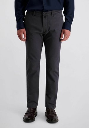 Mann trägt dunkelgraue Hose, dunkelbraune Lederschuhe und ein marineblaues Hemd mit Knöpfen und steht vor einem einfarbigen Hintergrund.