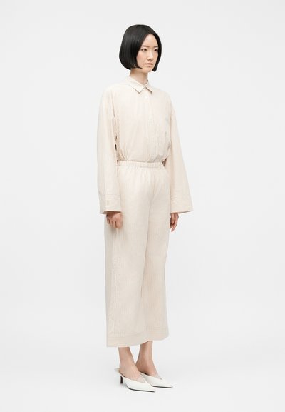 Γυναίκα με ίσια μαύρα μαλλιά φοράει κρεμ πουκαμίσα με ρίγες και μακριά μανίκια, παντελόνι φαρδύ στο ίδιο χρώμα και λευκές μυτερές γόβες, στέκεται προφίλ.