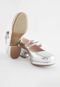 Silberne, metallische Mary Jane-Schuhe mit quadrater Zehenform, doppelten Riemen und niedrigem Blockabsatz. Die Laufsohle hat ein sternförmiges Profil.