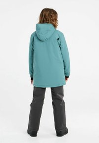 Türkise wasserdichte Jacke mit verstellbarer Kapuze, elastischen Bündchen und Logodetails, kombiniert mit grauen wasserdichten Hosen und schwarzen Stiefeln.