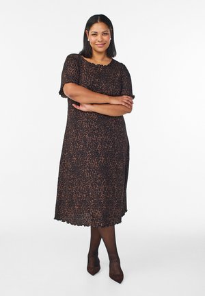 Ženska s temnimi, srednje dolgimi lasmi nosi rjavo črno obleko z leopardjim vzorcem, črne nogavice in temne pete, stoji z rokami prekrižanimi.