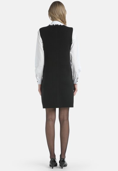 Abito nero senza maniche con taglio dritto, realizzato in tessuto morbido, abbinato a una camicia bianca con colletto arricciato e maniche lunghe.