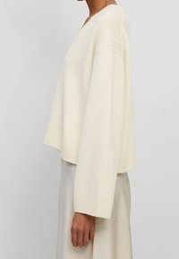 Pull blanc court en tissu tricoté doux. Il présente un décolleté en V profond, des manches larges et des poignets côtelés, offrant une coupe décontractée.