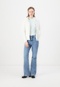 Giacca bianca imbottita con zip e cappuccio, maglietta azzurra chiara e jeans svasati blu. Tessuto liscio, vestibilità casual, hardware minimale. Stivali neri.