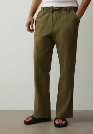 Osoba nosiaca olivovo zelené nohavice s elastickým pásom a sťahovacou šnúrou, kombinované s čiernymi sandálmi a bielym topom.