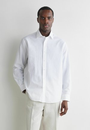 Mand stående mod en hvid væg iført en hvid skjorte med lange ærmer og lyse bukser, med den ene hånd i lommen.