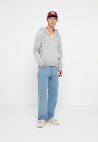 Grauer Zip-Hoodie mit Kordelzügen, kombiniert mit hellblauen weitgeschnittenen Jeans und weißen Sneakern. Das Model trägt eine burgunderfarbene Mütze.