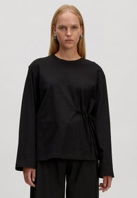 Zwarte langemouwen top van zacht materiaal, met een zijstrikdetail voor een geraffineerde uitstraling en een ontspannen pasvorm. Ronde halslijn, zonder patronen.