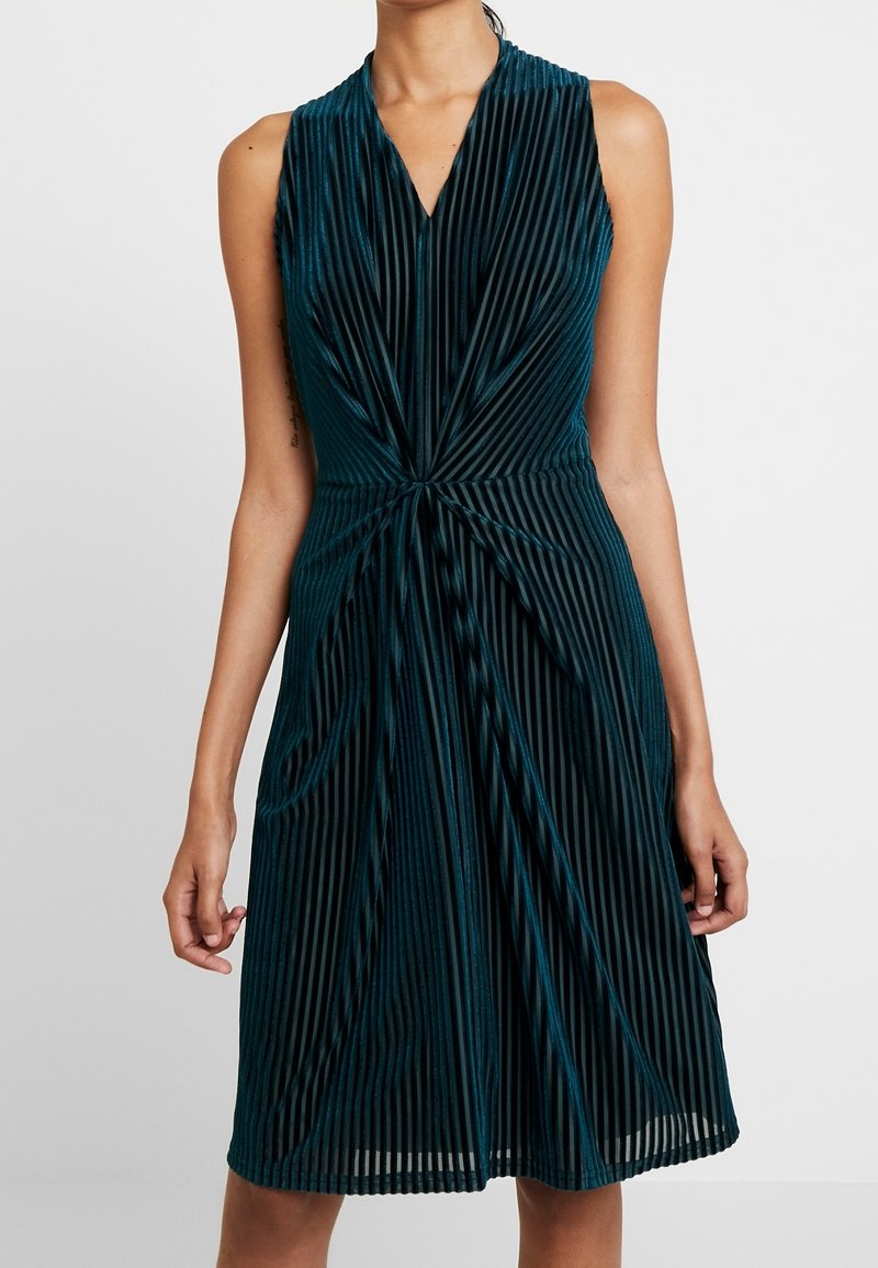 Robe sans manches en velours vert foncé avec des rayures verticales et un détail en tissu torsadé à la taille, longueur genou, portée par une personne.