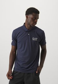 Navy polo shirt made of smooth, breathable fabric. Features a collar, short sleeves, and an EA7 logo embroidered on the chest.